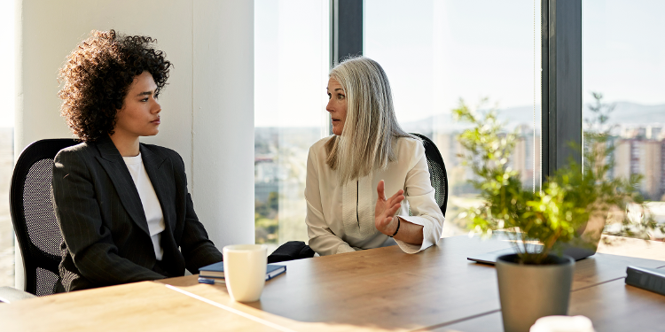 Two professional women sit at a conference table in conversation as one gestures with her hand